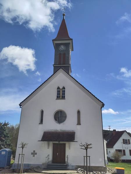 Quelle: Sybille Konstanzer, Röm.-kath. Kirchengemeinde Sigmaringen KdöR Außenansicht der Kirche St. Fidelis in Otterswang von vorne. Weiße Kirche mit dunklem Kirchturm. Im Hintergrund blauer Himmel mit weißen Wolken.