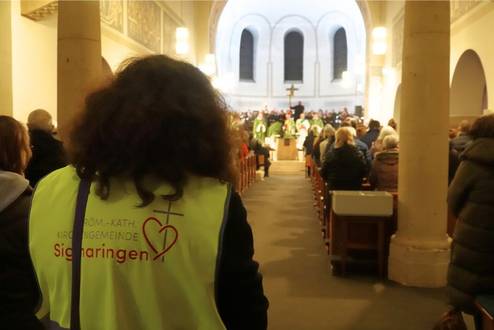 Eine Ordnerin in einer gelben Warnweste steht mit dem Rücken zur Kamera. Im Hintergrund findet ein Gottesdienst statt.