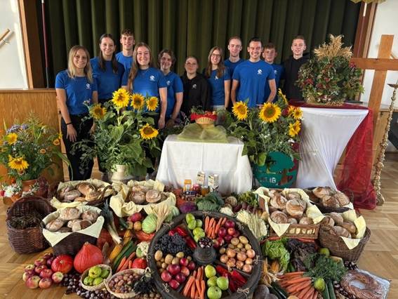 Auf dem Bild ist ein bunter Erntedankaltar mit verschiedenem Obst und Gemüse, Broten und Sonnenblumen zu sehen. Im Hintergrund stehen Jugendliche mit blauem T-Shirt. Der Altar befindet sich vor einer Bühne.